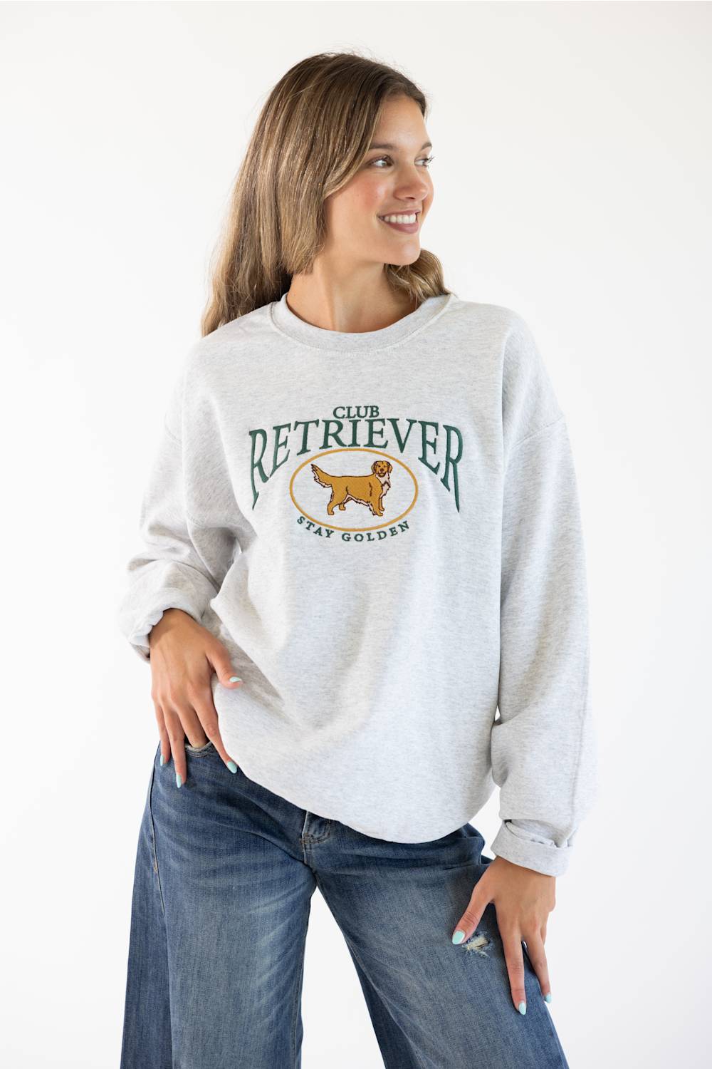 Front ¾ view of a female model wearing the Sweet Claire Embroidered Club Retriever Sweatshirt in Ash heather grey, featuring a green "Club Retriever Stay Golden" chest embroidery, crew neck, and relaxed fit paired with denim jeans.