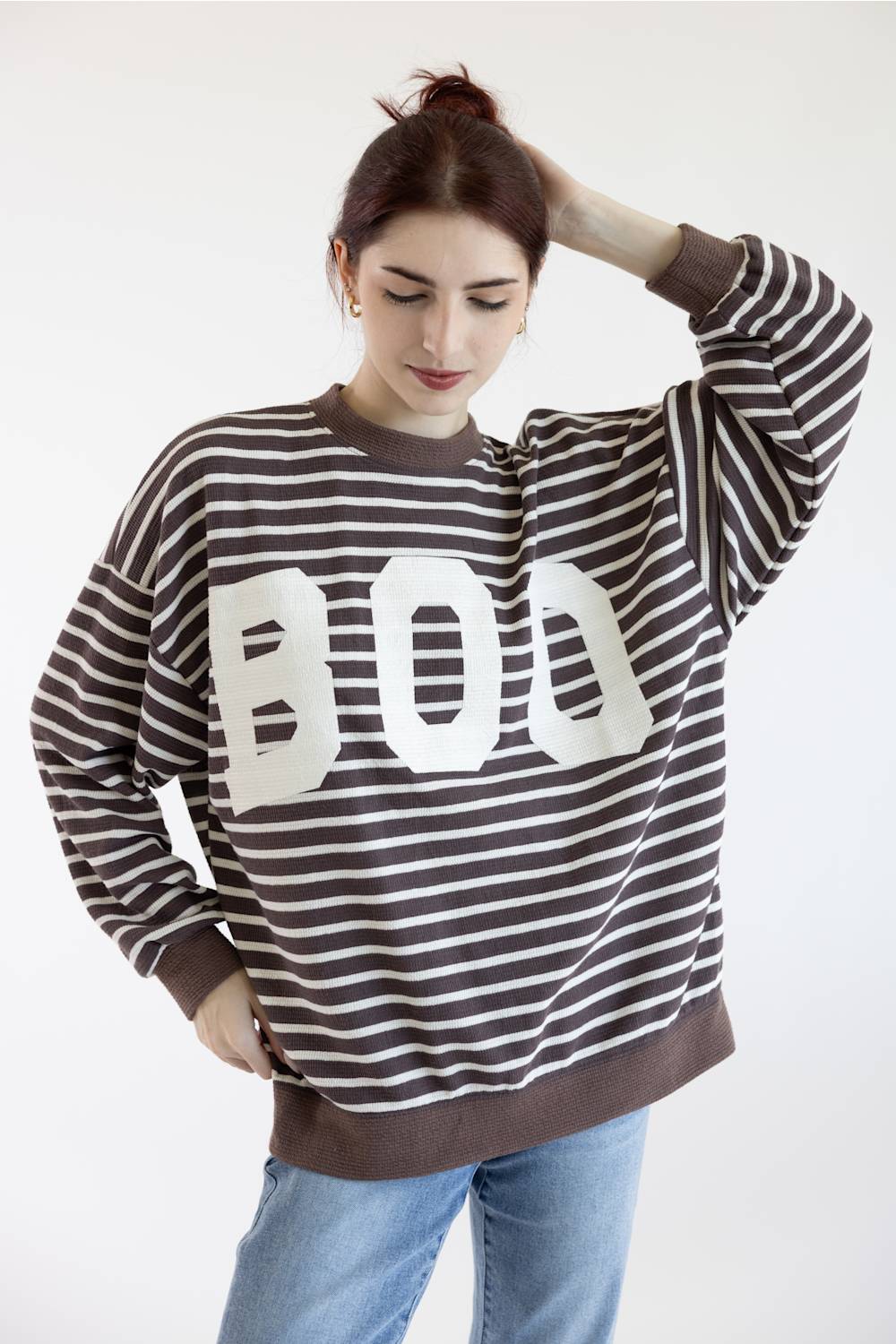 A woman posing in a brown and white horizontally striped sweatshirt with a large white BOO graphic on the front, paired with light blue jeans, against a plain white background.