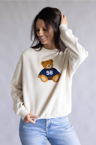 A woman wearing a cream-colored crewneck sweatshirt with a teddy bear graphic on the front, paired with light blue jeans, posing against a neutral gray studio background.