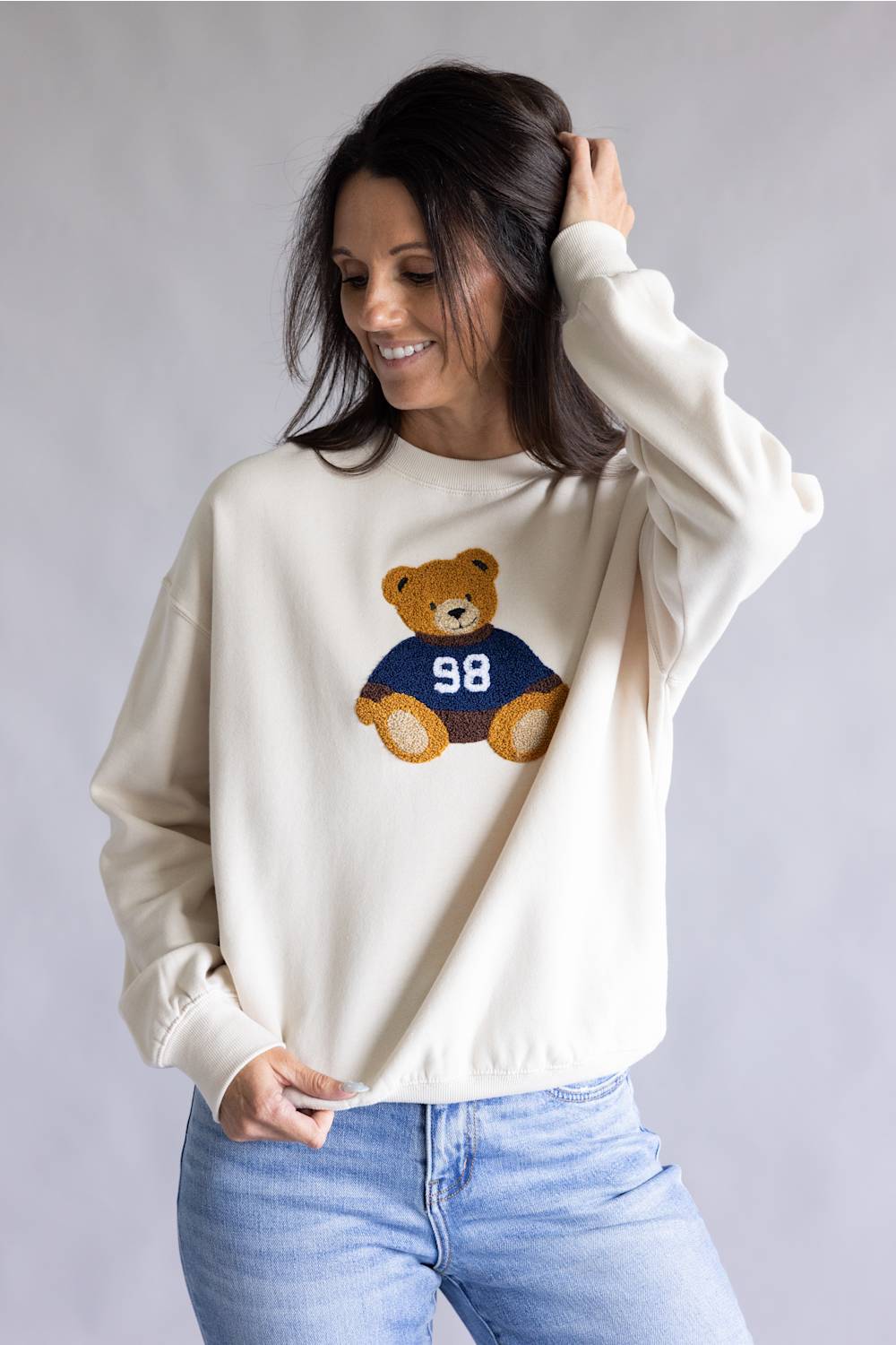 A woman wearing a cream-colored crewneck sweatshirt with a teddy bear graphic on the front, paired with light blue jeans, posing against a neutral gray studio background.
