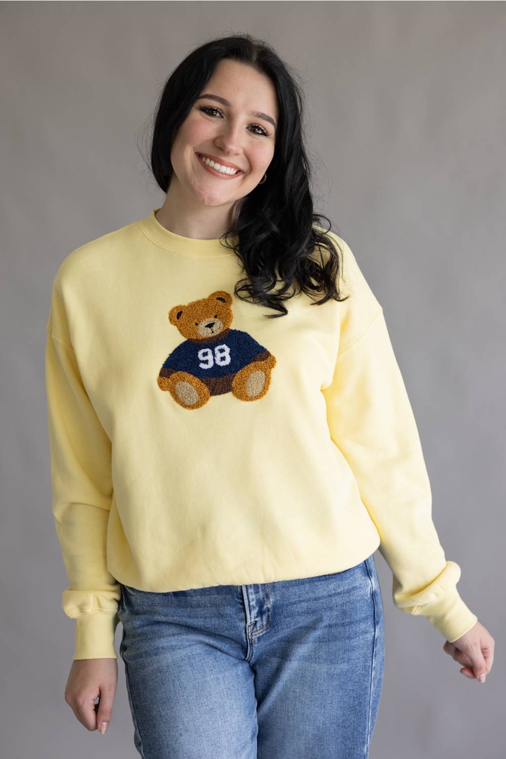 A smiling woman wearing a pale yellow crewneck sweatshirt with a teddy bear graphic wearing a navy blue jersey with the number 98, paired with blue jeans, against a neutral gray background.