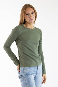 A woman modeling a long-sleeve crewneck knit top in olive green, paired with light-wash blue jeans, against a plain white studio background.