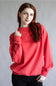 A woman with long red hair wearing a bright coral-red crewneck sweatshirt and black pants, posing against a neutral gray studio background.