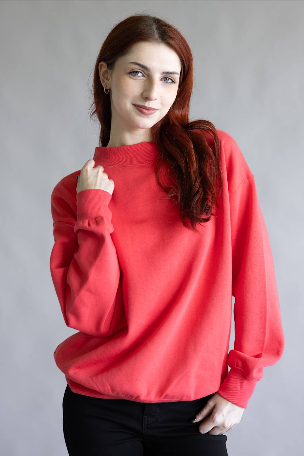 A woman with long red hair wearing a bright coral-red crewneck sweatshirt and black pants, posing against a neutral gray studio background.