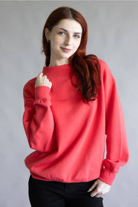 A woman with long red hair wearing a bright coral-red crewneck sweatshirt and black pants, posing against a neutral gray studio background.
