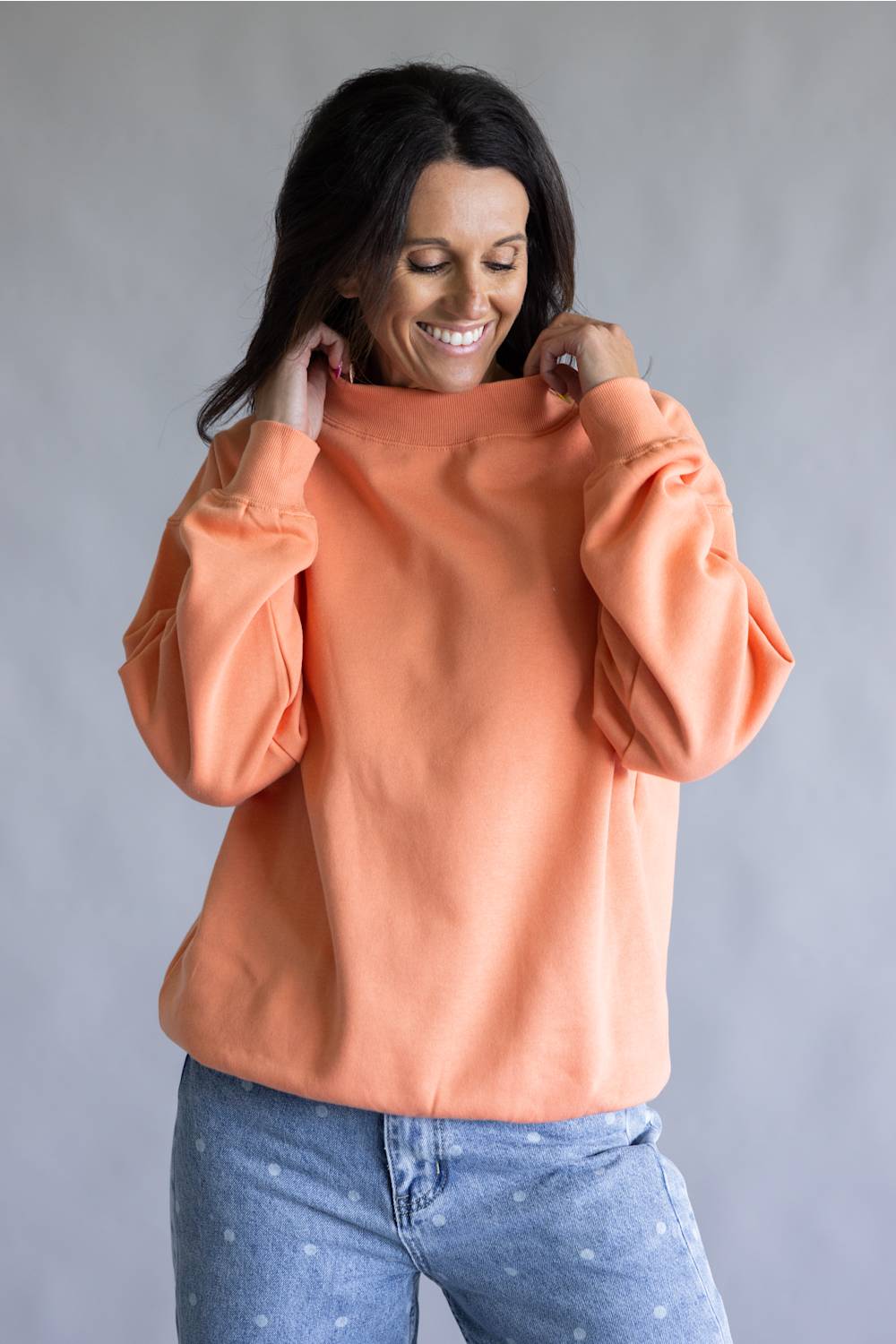 A woman wearing a peach-colored crewneck sweatshirt and light blue jeans, smiling and pulling the collar of the sweatshirt up slightly.