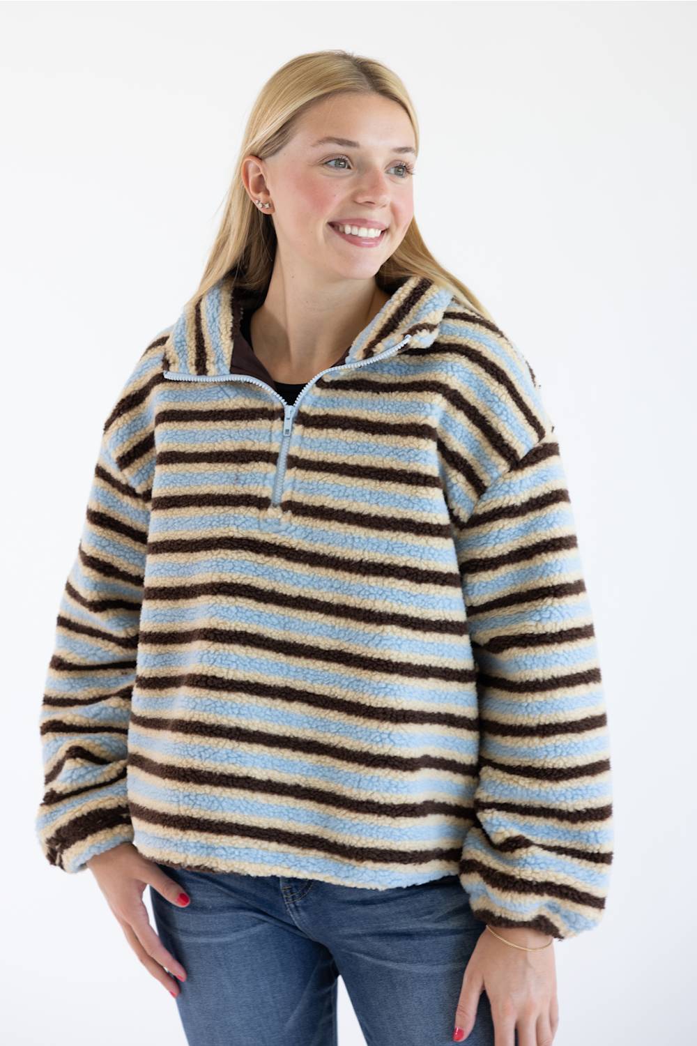A woman wearing a fuzzy quarter-zip pullover sweater with horizontal brown, light blue, and cream stripes, paired with blue jeans, against a plain white background.