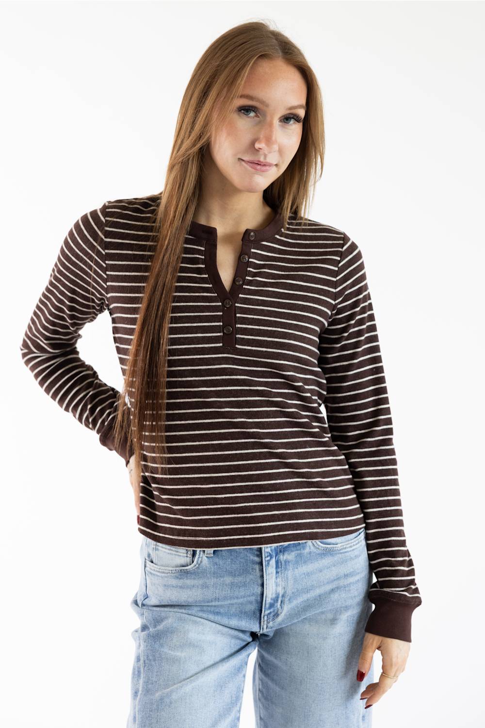 A woman modeling a brown and white horizontally striped long-sleeve henley shirt with a split button placket, paired with light blue jeans, against a plain white background.