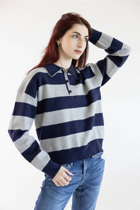 A woman wearing a navy and light gray horizontally striped long-sleeve polo sweater with blue jeans, posing against a plain white studio background.