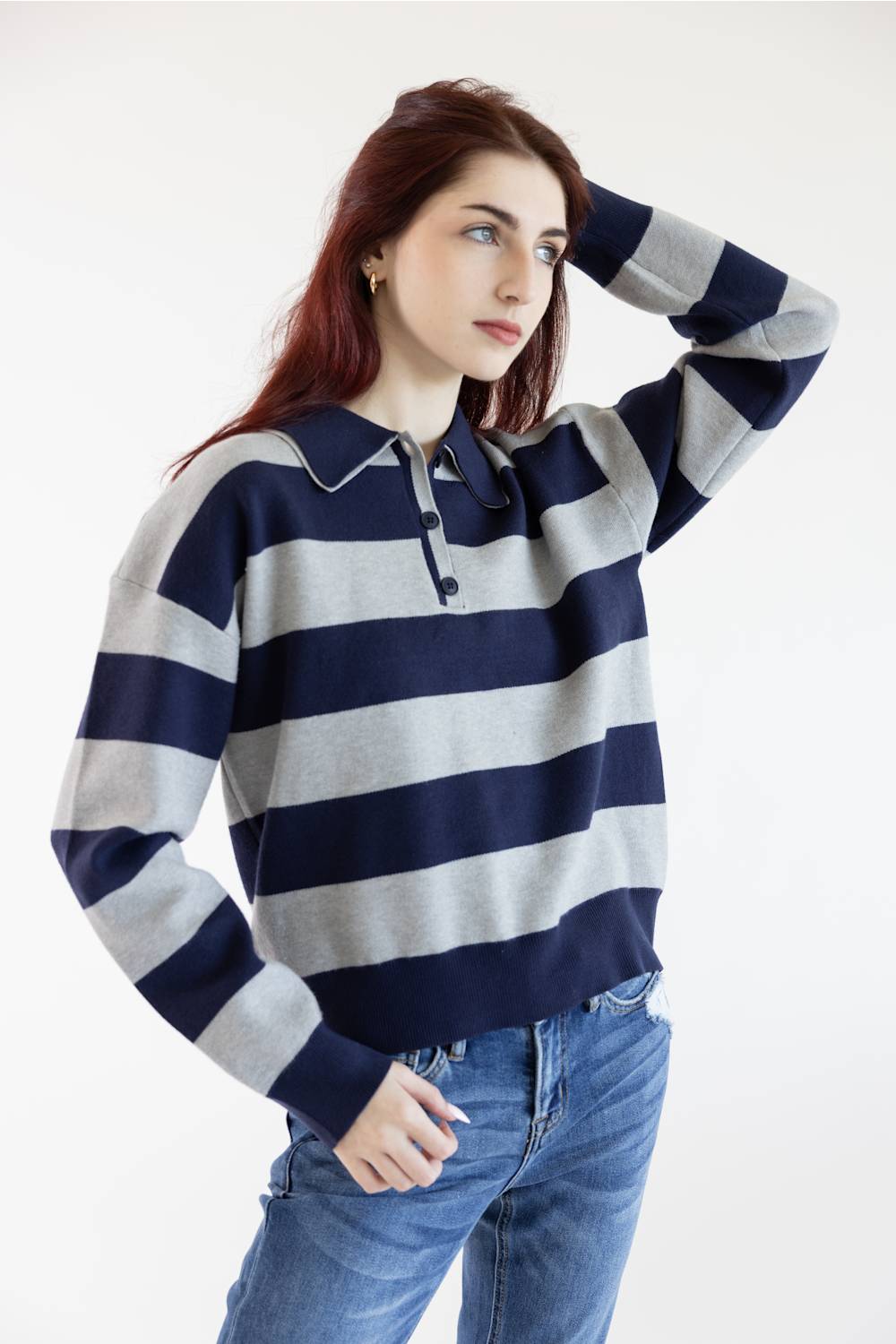 A woman wearing a navy and light gray horizontally striped long-sleeve polo sweater with blue jeans, posing against a plain white studio background.