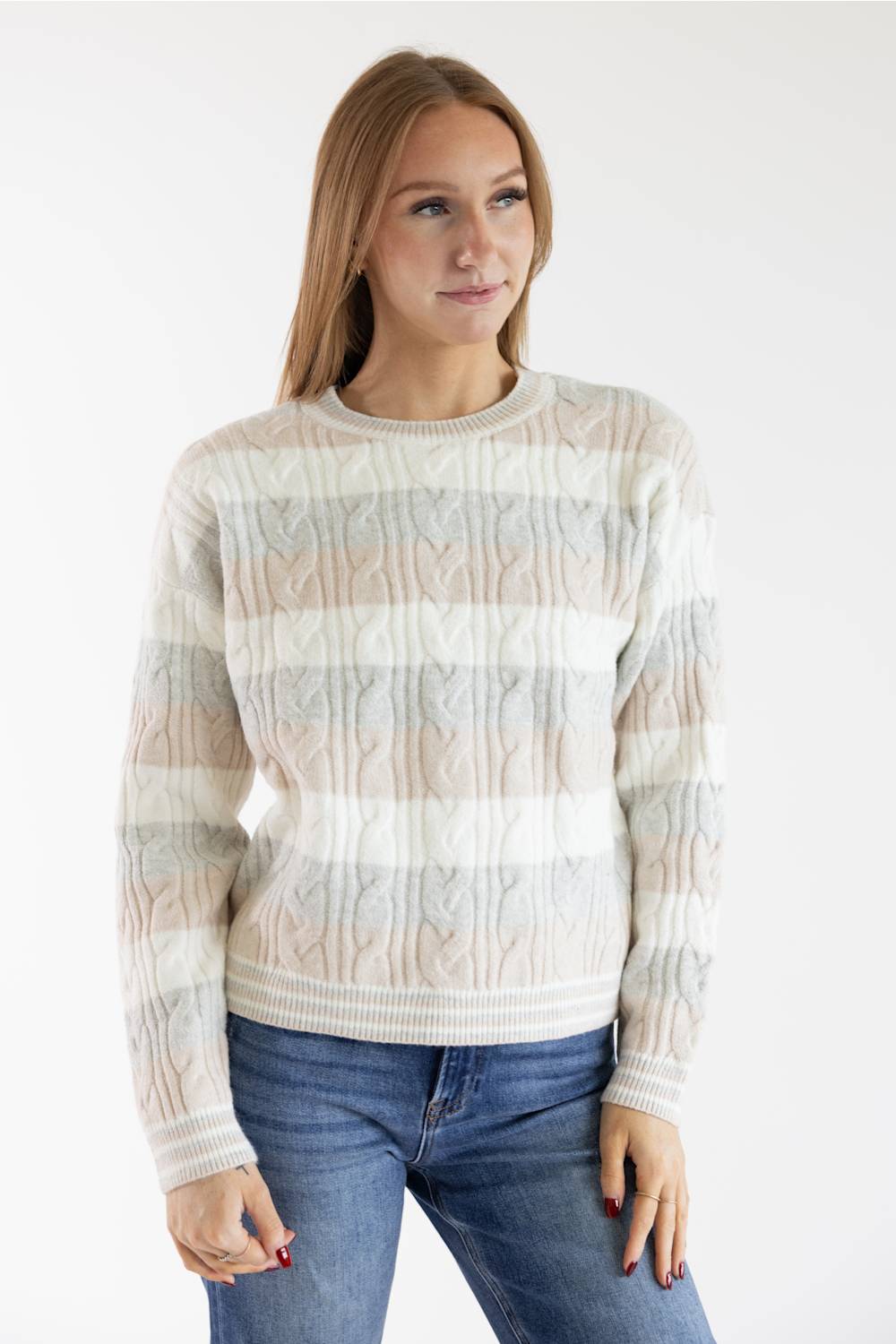 A woman wearing a cream, beige, and light gray striped cable-knit sweater with blue jeans, standing against a plain white studio background.