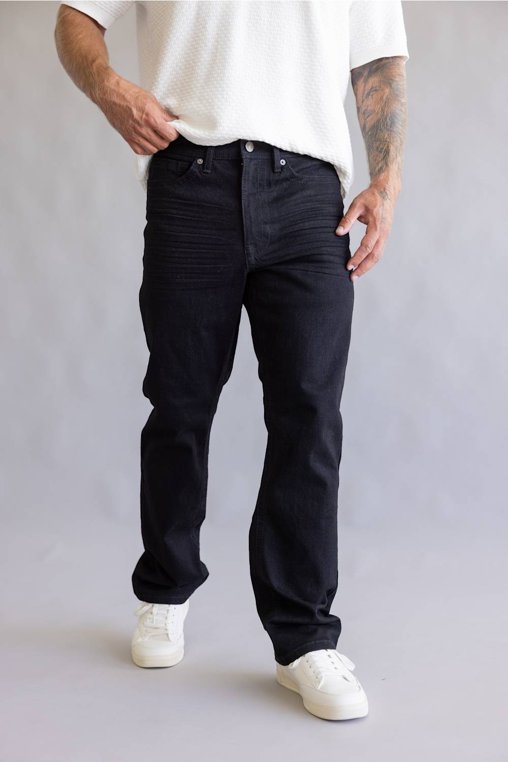 A person wearing black slim-fit jeans with a textured vertical stripe pattern, styled with a white t-shirt and white sneakers against a light gray studio background.