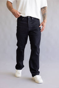 A person wearing black slim-fit jeans with a textured vertical stripe pattern, styled with a white t-shirt and white sneakers against a light gray studio background.