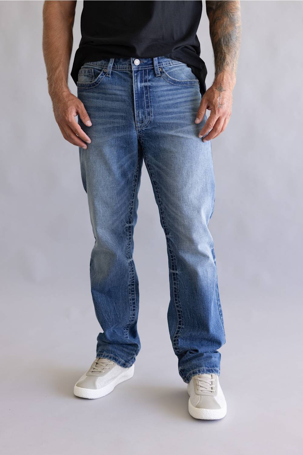 A person wearing light-to-medium wash blue jeans, a black t-shirt, and white sneakers, shown from the waist down against a plain light gray studio background.
