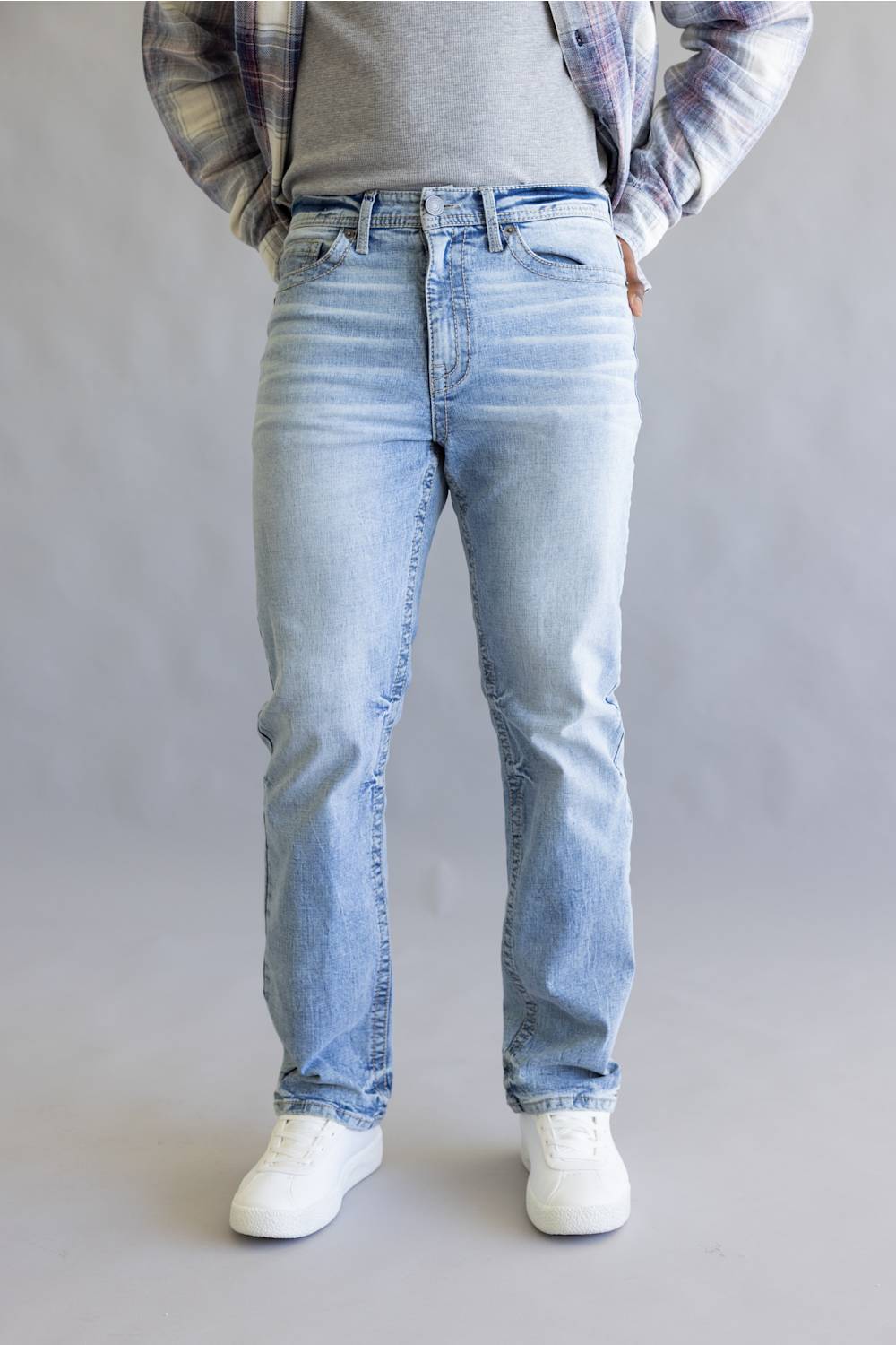 A person wearing light-wash blue denim jeans, a gray t-shirt, a plaid flannel shirt, and white sneakers, standing against a neutral gray studio background.