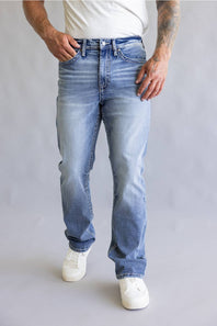 Rear view of a person wearing light blue denim jeans with decorative embroidered patch detailing on the back pockets, paired with a white t-shirt against a neutral gray background.