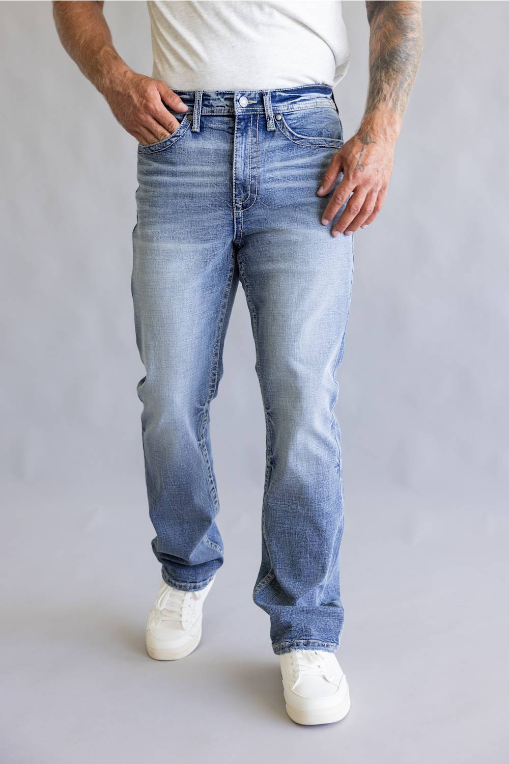 Rear view of a person wearing light blue denim jeans with decorative embroidered patch detailing on the back pockets, paired with a white t-shirt against a neutral gray background.