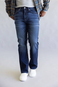 A person wearing dark blue straight-leg denim jeans, styled with a gray t-shirt, a blue and brown plaid flannel shirt, and white sneakers, standing against a light gray studio background.