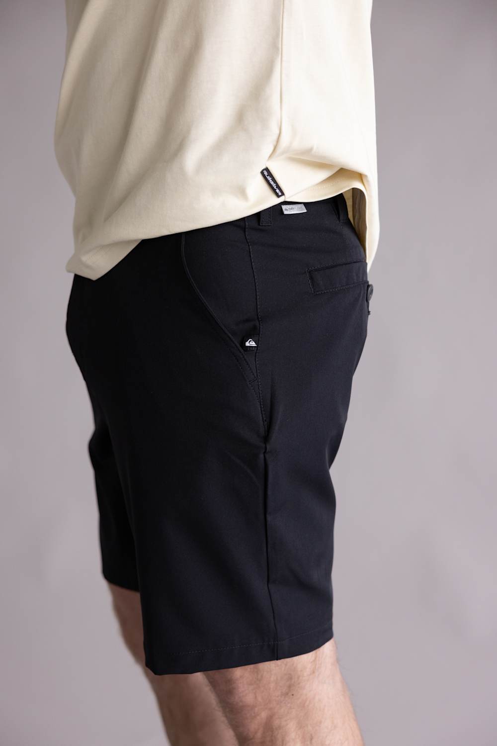 Side view of a person wearing black shorts with a plain background. The shorts are the Quiksilver Taxer Amphibian 18" Hybrid Shorts for Men in Black