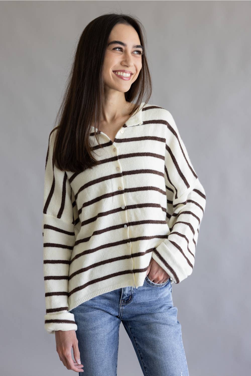 A woman wearing a cream knit button-up shirt with dark brown horizontal stripes and blue jeans, standing against a neutral gray studio background.