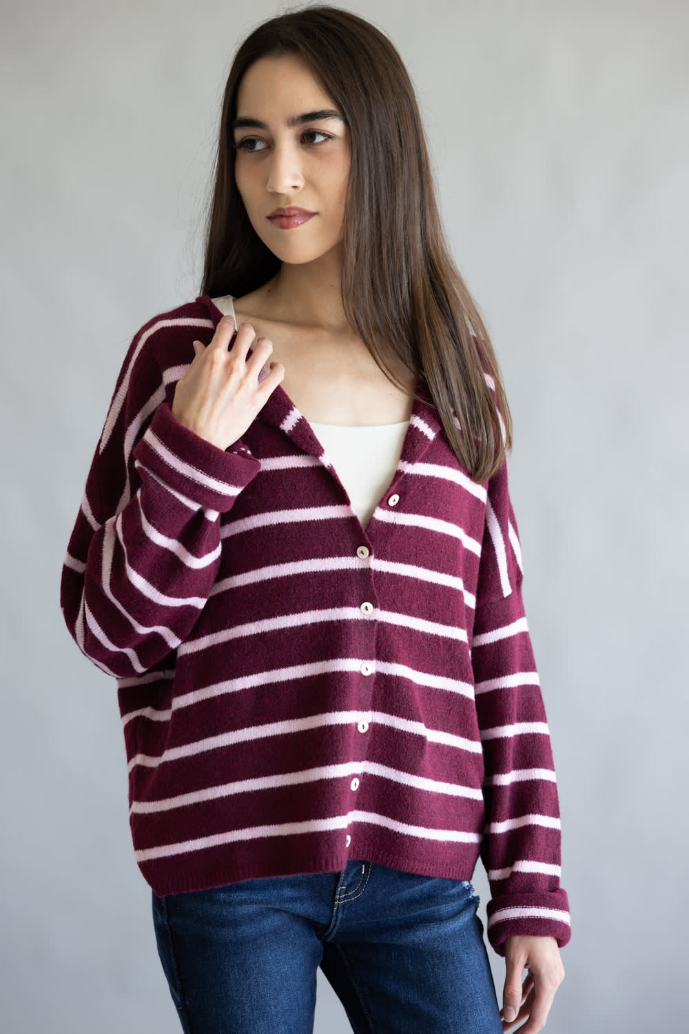 A woman wearing a burgundy and light pink striped knit cardigan over a white top and blue jeans, standing against a plain gray background.