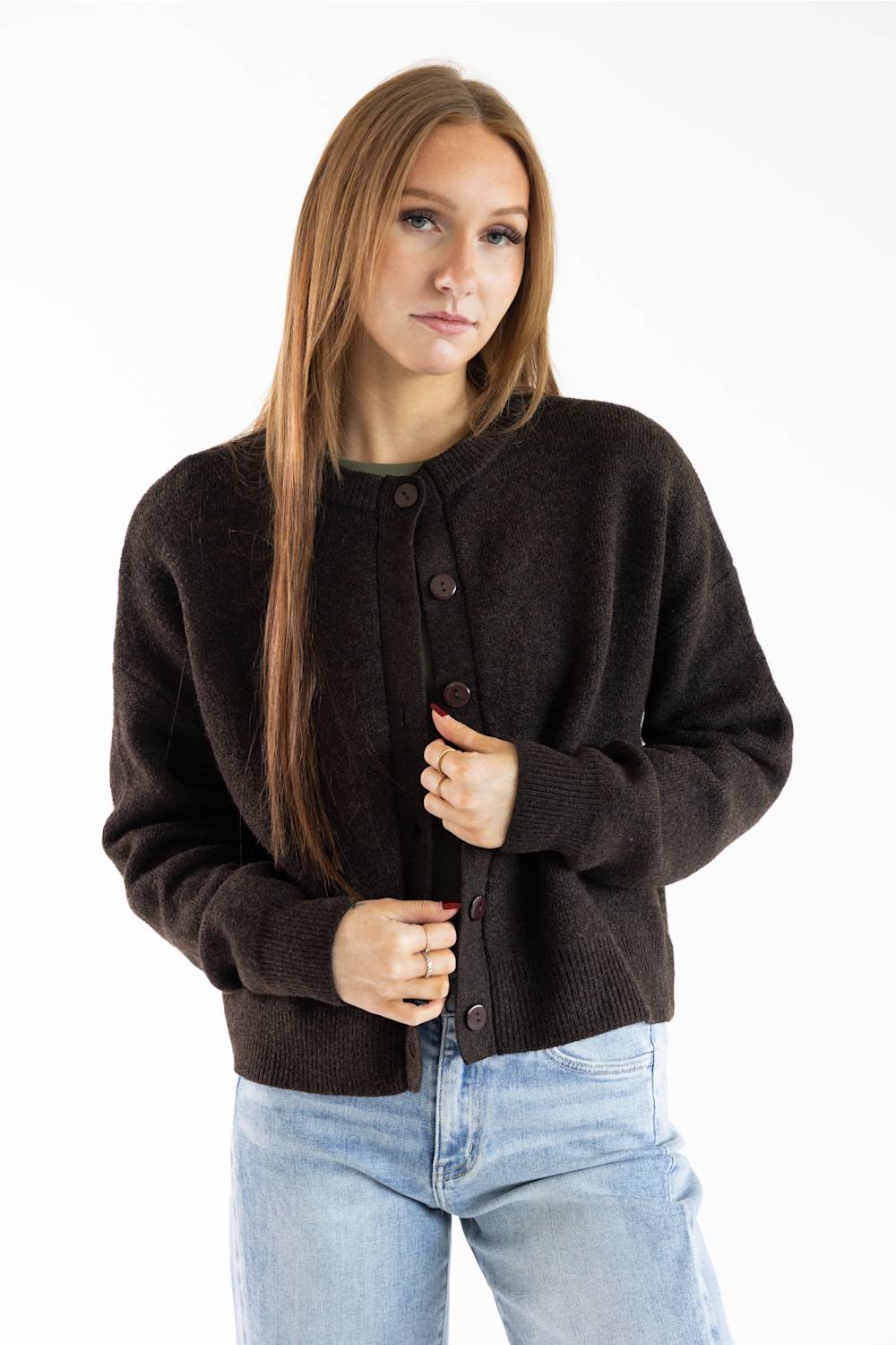 A woman with long light brown hair wearing a dark brown button-up cardigan sweater and light blue jeans, standing against a plain white studio background.