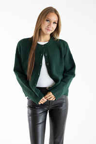 A woman modeling a dark green cropped button-up cardigan over a white top and black faux-leather pants, photographed against a plain white studio background.