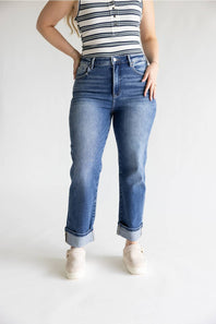 A model wearing a pair of high-waisted, straight-leg blue jeans with rolled cuffs, paired with a striped tank top and white sneakers, photographed against a plain white studio background.