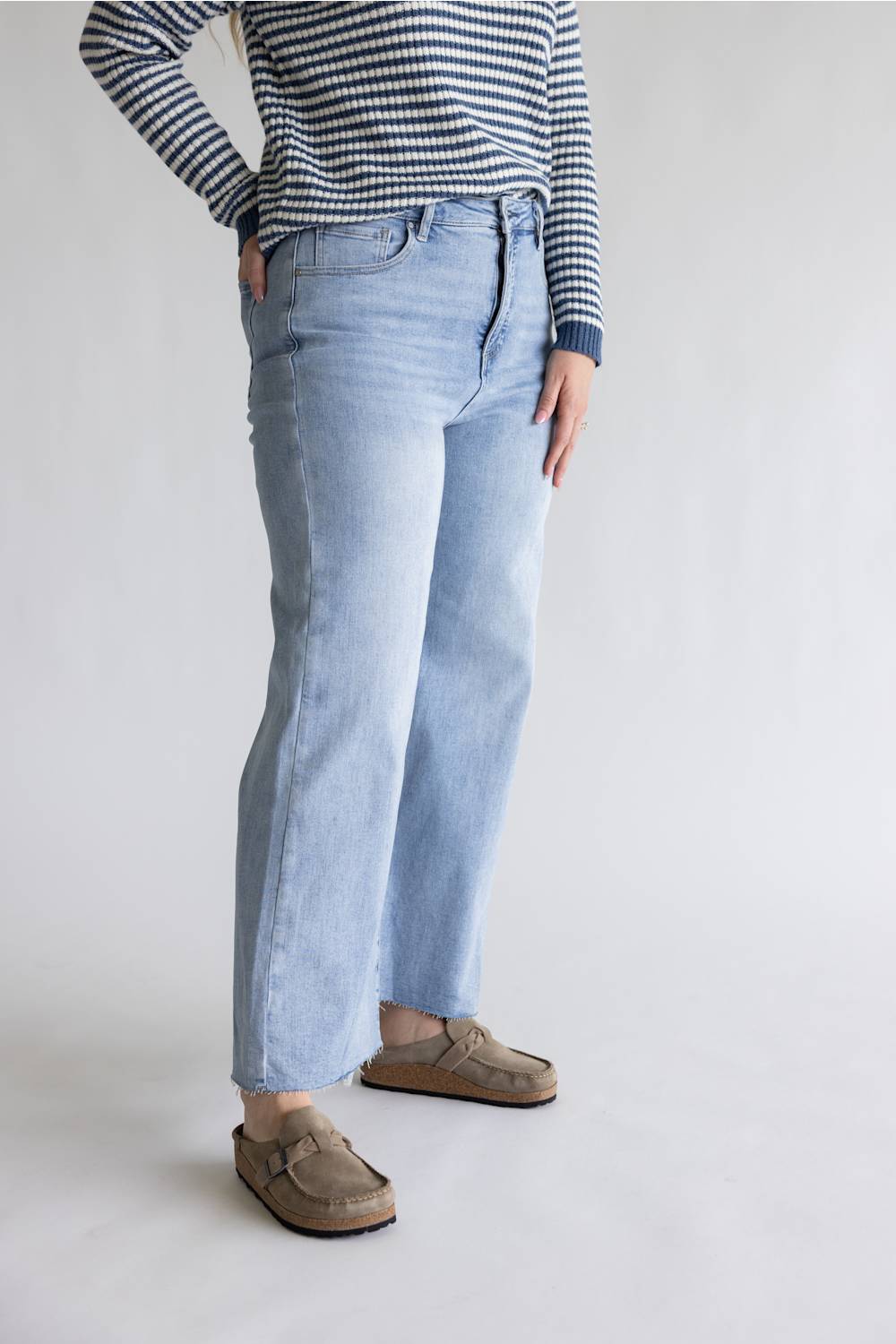 A person wearing light-wash blue straight-leg jeans, a navy and white striped long-sleeve top, and brown suede loafers, standing against a plain light gray studio background.