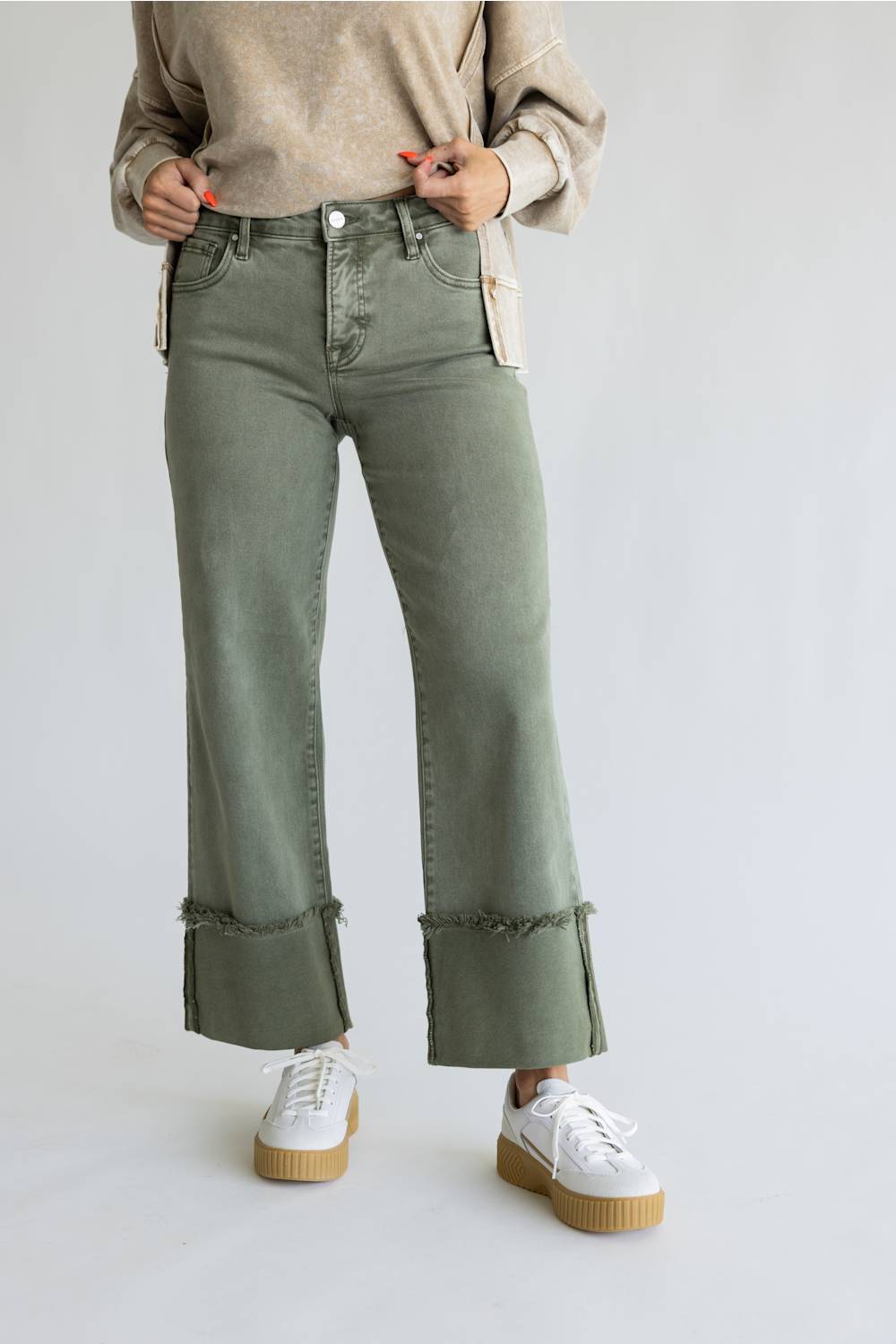 A person wearing olive green wide-leg cropped jeans with rolled frayed hems, styled with a beige sweater and white platform sneakers against a light gray studio background.