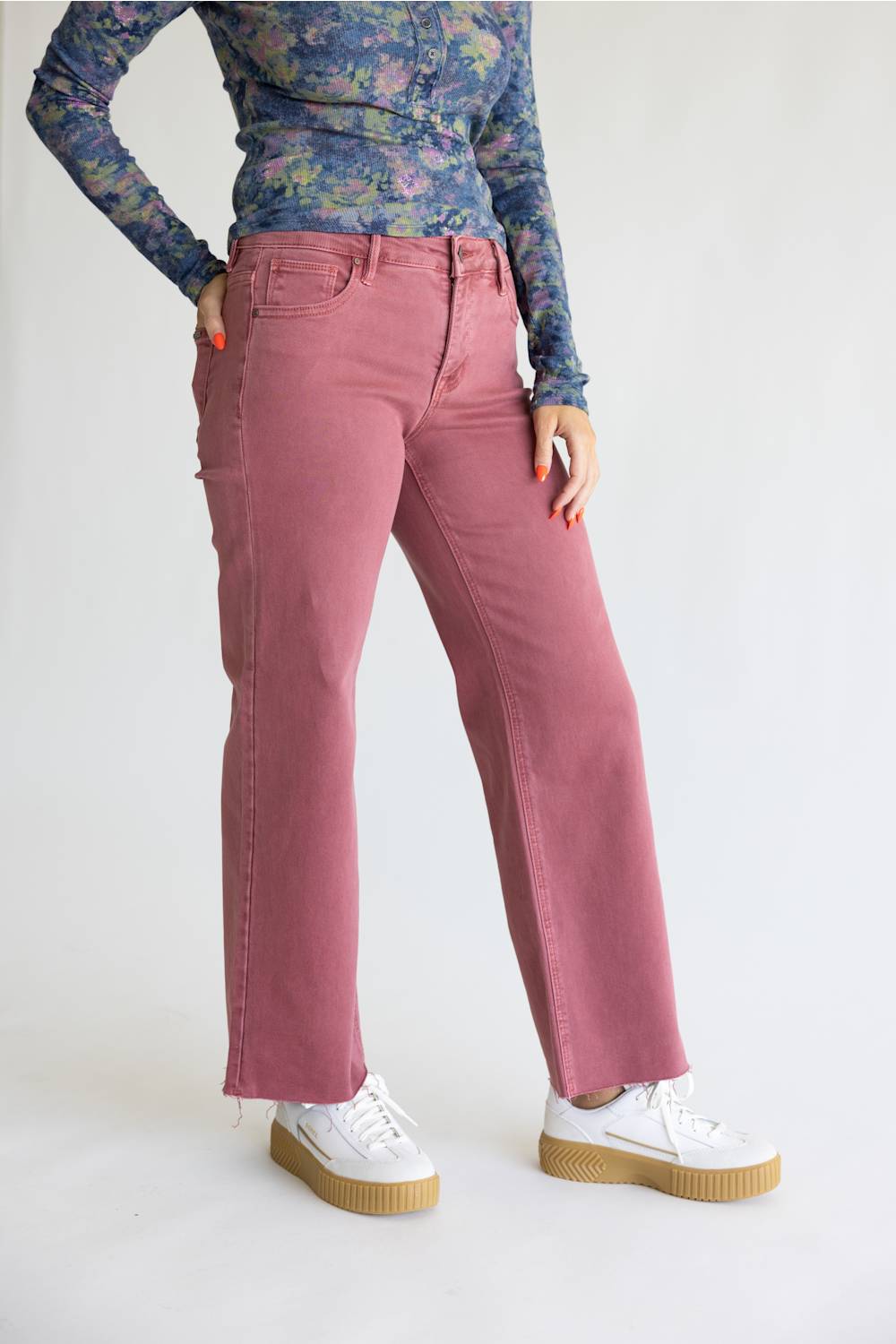 A model wearing a pair of dusty rose straight-leg jeans, styled with a blue floral long-sleeve top and white platform sneakers, photographed against a plain white studio background.