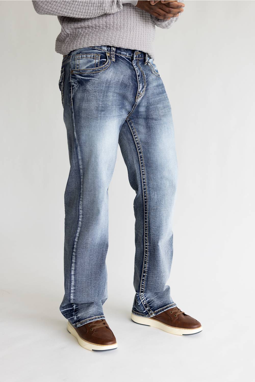 A person wearing light blue faded denim jeans with brown slip-on shoes and a gray knit sweater, standing against a plain light gray studio background.