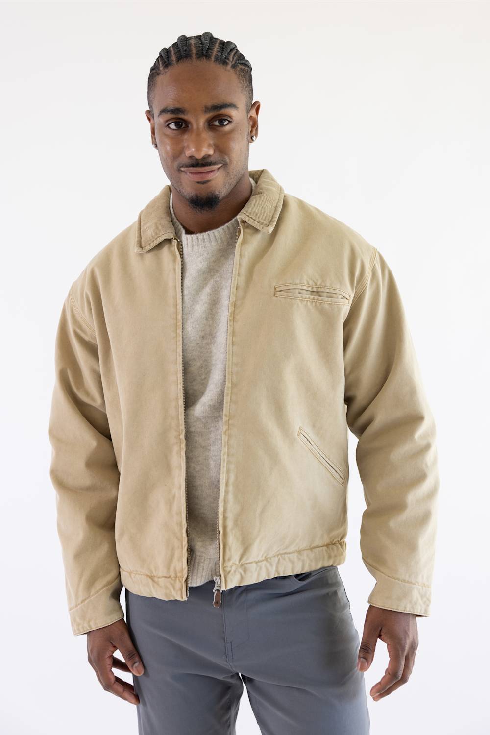 A man wearing a beige utility jacket over a light gray sweater and gray pants, standing against a plain white studio background.