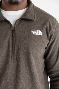 A close-up of a person wearing a brown textured quarter-zip fleece pullover by The North Face, layered over a white t-shirt. The brand logo is visible on the left chest.
