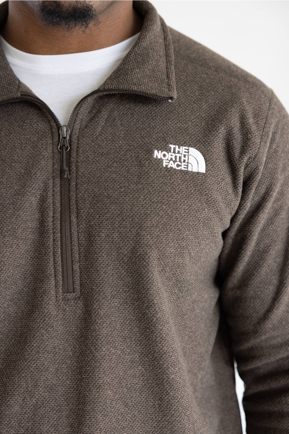 A close-up of a person wearing a brown textured quarter-zip fleece pullover by The North Face, layered over a white t-shirt. The brand logo is visible on the left chest.