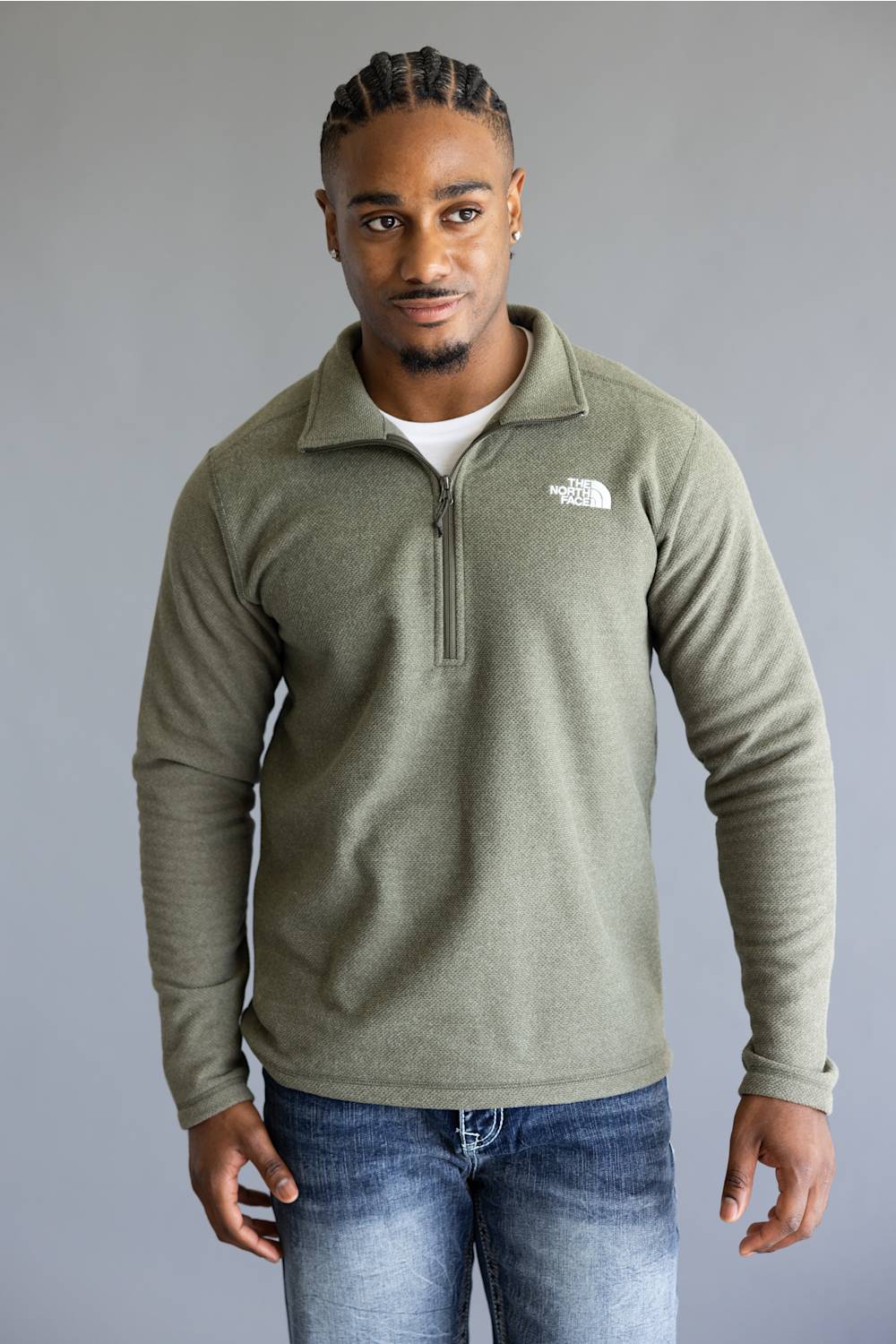 A man wearing an olive green quarter-zip fleece pullover by The North Face, paired with blue jeans, standing against a neutral gray studio background.