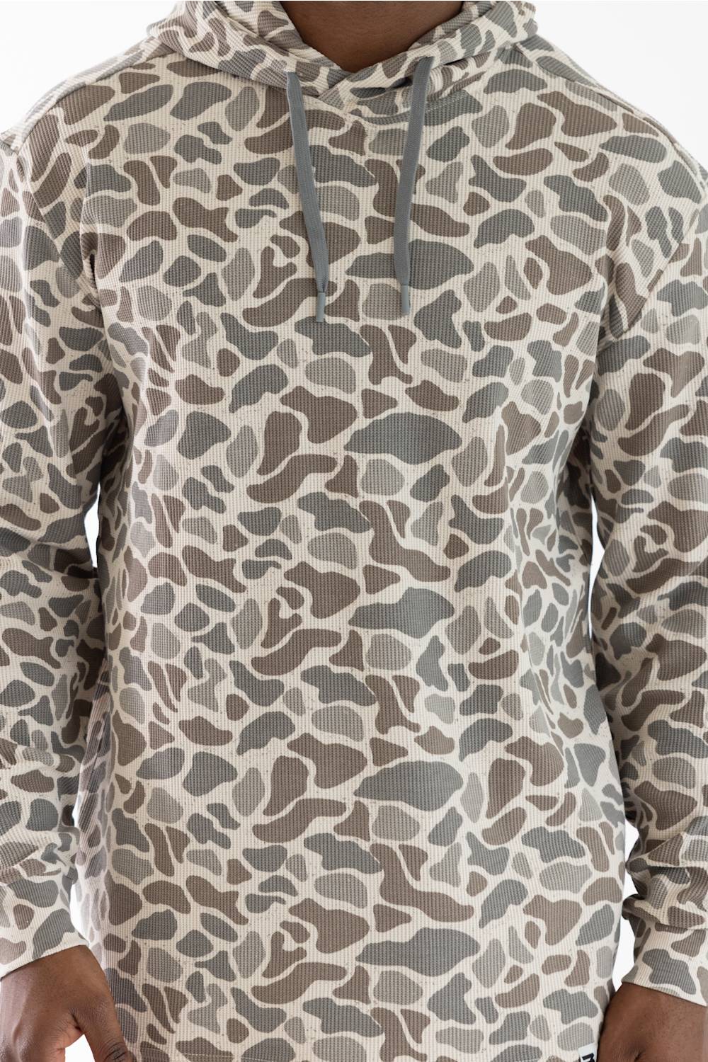 A close-up of a long-sleeve hooded shirt with a beige, brown, and gray organic camo print, featuring gray drawstrings.