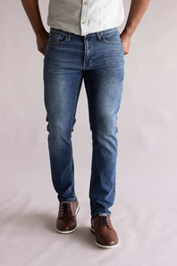 Person wearing blue jeans and brown shoes on a plain background. The jeans are Weatherproof Vintage Slim Jeans for Men in Tidepool Tint.