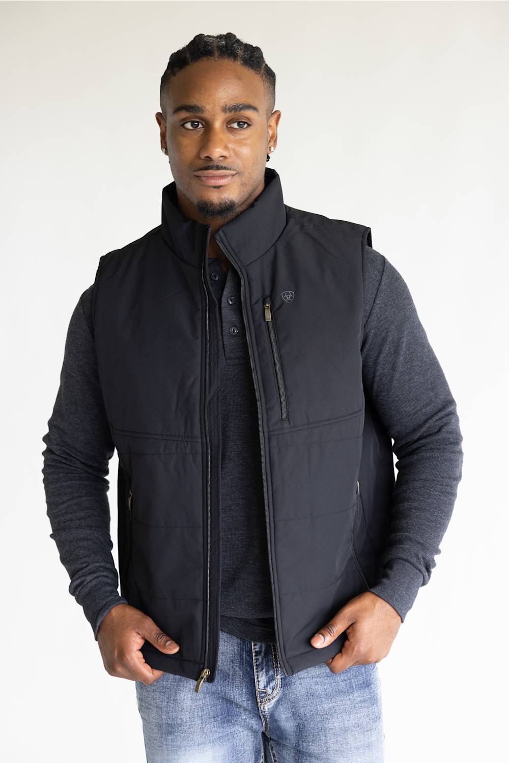 A man wearing a black zip-up sleeveless vest over a dark gray long-sleeve shirt and blue jeans, standing against a plain white studio background.