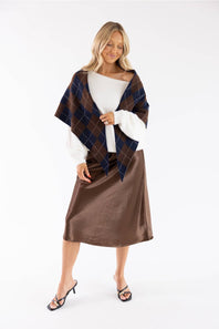 A woman modeling a brown and navy blue argyle plaid wrap shawl draped over a white off-the-shoulder long-sleeve top and a brown satin midi skirt, paired with black strappy heels, against a plain white studio background.