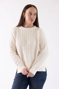 A woman wearing a cream-colored, textured cable-knit sweater and dark blue jeans, standing against a plain white studio background.