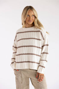 A woman wearing a cream ribbed knit sweater with brown horizontal stripes and beige cargo pants, photographed against a plain light gray studio background.