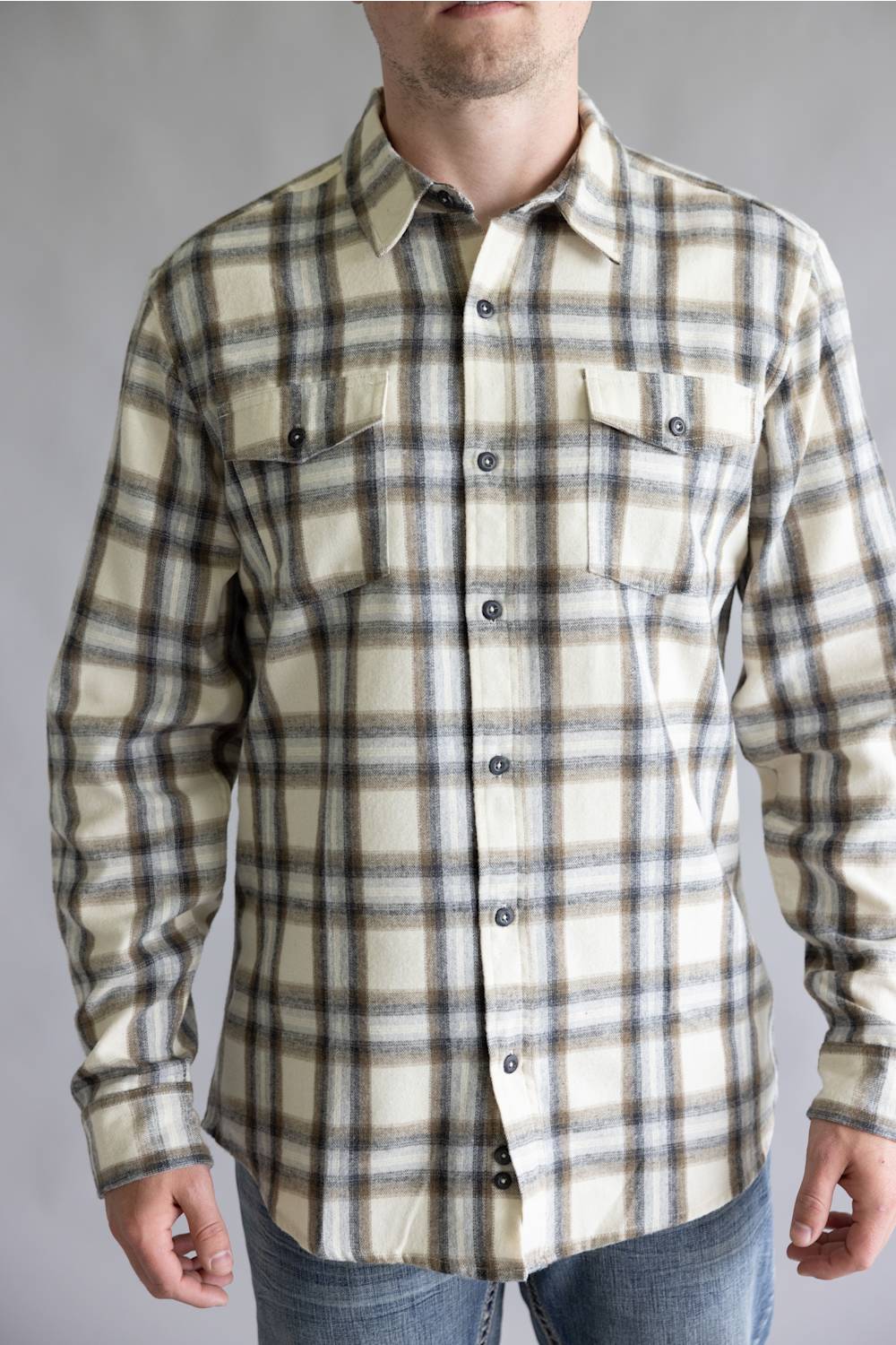 Close-up front view of a male model wearing the Split Flannel Shirt for Men in Grey Ecru, showcasing the beige, gray, and brown plaid pattern, dual buttoned flap chest pockets, long sleeves, and button-down front against a neutral gray background.