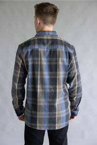 Back view of a male model wearing the Split Flannel Shirt for Men in Moka, showcasing the blue, dark gray, and tan plaid pattern across the back, straight yoke, long sleeves, and rounded hem against a neutral gray background.