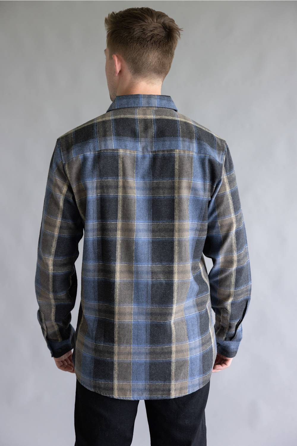Back view of a male model wearing the Split Flannel Shirt for Men in Moka, showcasing the blue, dark gray, and tan plaid pattern across the back, straight yoke, long sleeves, and rounded hem against a neutral gray background.