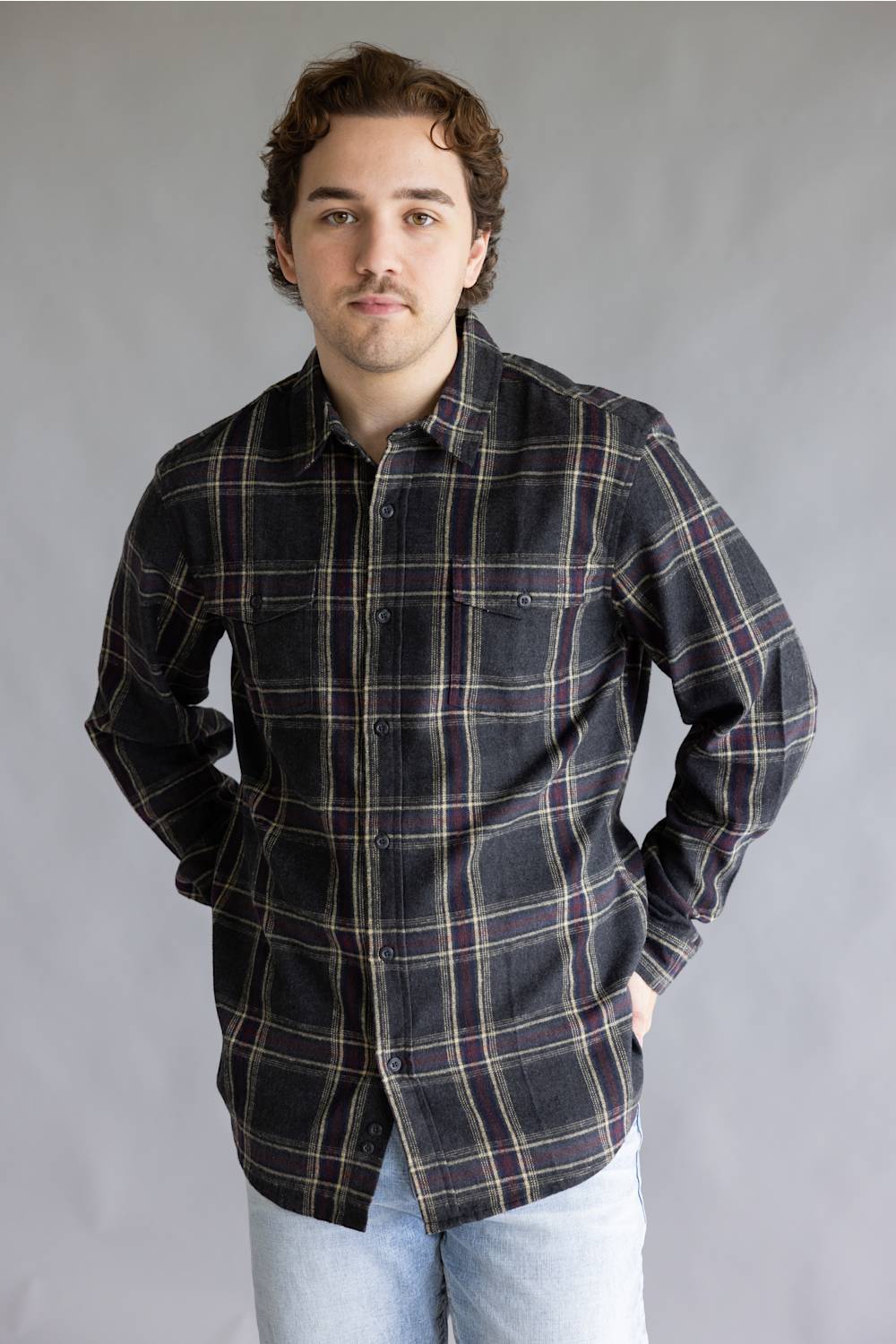 Front view of a male model with hands in his pockets wearing the Split Flannel Shirt for Men in Slate, showcasing the charcoal gray, burgundy, and cream plaid pattern, long sleeves, and double chest button pockets against a gray background.