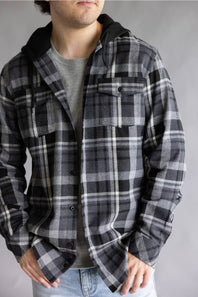 Close-up front view of a model wearing an open, unbuttoned Split Men's long-sleeve hooded flannel shirt in a black, grey, and white plaid pattern, styled over a grey ribbed shirt with hands in blue jean pockets against a grey studio background.