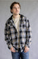 Model wearing the Split Men's long-sleeve hooded flannel shirt in a cream, black, and tan plaid pattern, featuring two chest flap pockets and an attached black hood with drawstrings against a gray studio background.