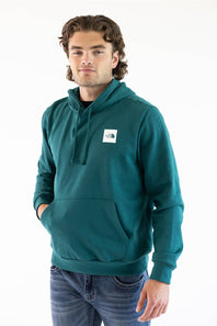 A man wearing a teal green pullover hoodie with a white square The North Face logo patch on the left chest, paired with blue jeans, photographed against a plain white studio background.