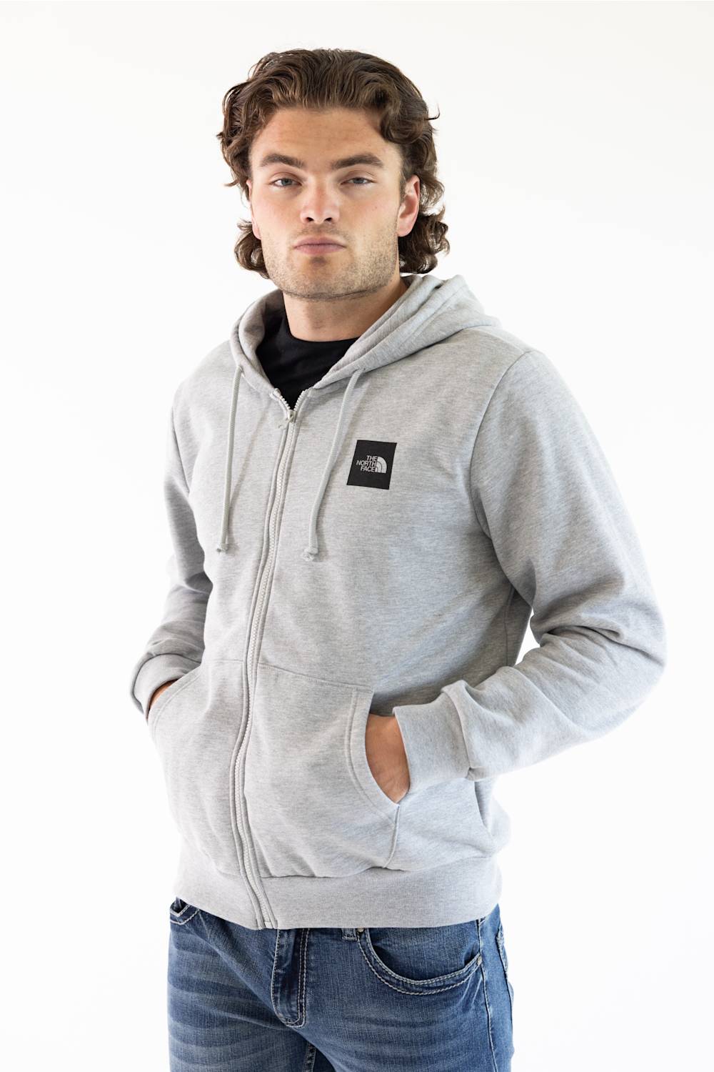 A man wearing a light gray zip-up hoodie with a black The North Face logo patch on the chest, paired with blue jeans, photographed against a plain white background.